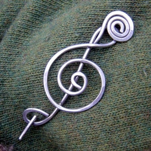 May include: A silver treble clef shaped brooch with a spiral design. The brooch is pinned to a green knitted fabric.