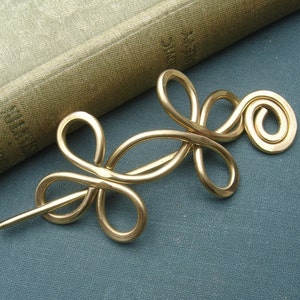 May include: A gold metal hair pin with a spiral design and a knot in the center. The pin is on a blue surface.