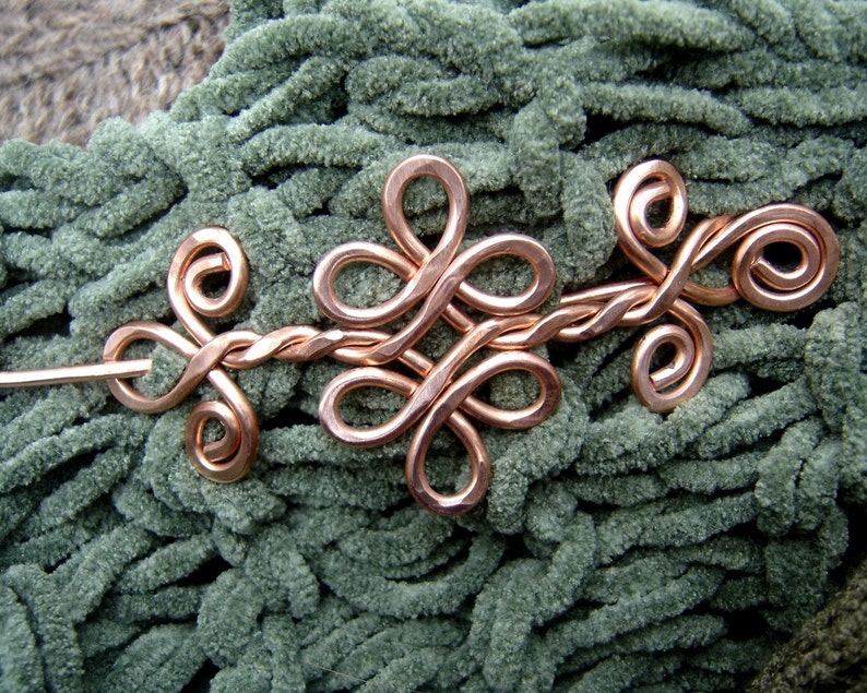 May include: A copper wire hair pin with a decorative knot design. The pin is on a green fuzzy surface.