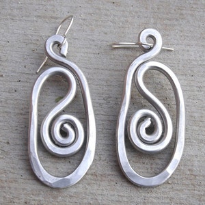 May include: A pair of silver earrings with a unique design. Each earring features a large oval shape with a smaller spiral inside. The earrings are made of metal and have a polished finish.