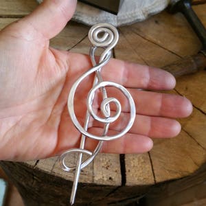 May include: A silver treble clef hair pin, handcrafted from wire, with a hammered finish. The pin is shaped like a treble clef with a spiral at the top.