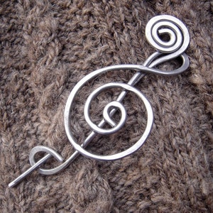 May include: A silver metal treble clef brooch with a spiral design. The brooch is on a brown knitted fabric.