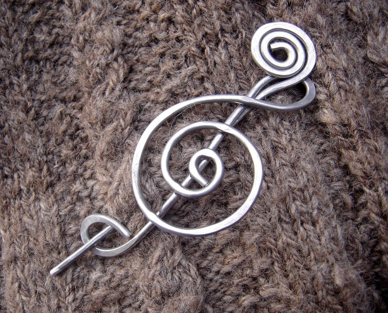 May include: A silver metal treble clef brooch with a spiral design. The brooch is on a brown knitted fabric.