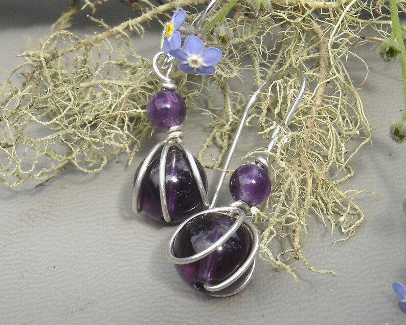 May include: A pair of silver earrings with purple amethyst stones. The stones are encased in a wire cage design.