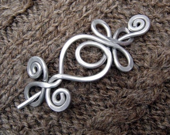 May include: A silver metal brooch with a swirling, abstract design. The brooch is on a brown knitted fabric.