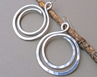 May include: A pair of silver hoop earrings with a hammered finish. The earrings are made of two circles, one inside the other, and are connected by a small loop at the top.