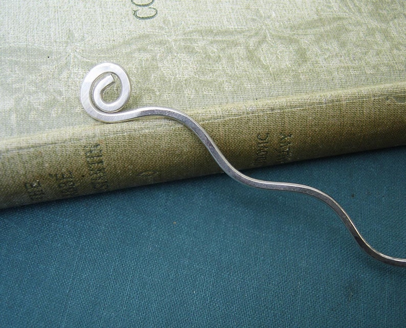 May include: A silver bookmark with a curled end. The bookmark is laying on a green book and a blue surface.