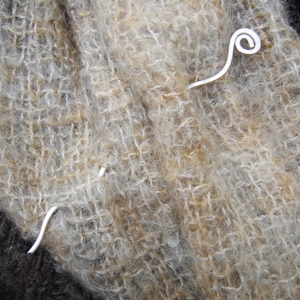 May include: A silver metal shawl pin with a curled end. The pin is resting on a brown and white fuzzy fabric.