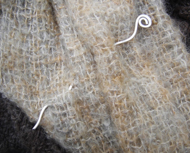 May include: A silver metal shawl pin with a curled end. The pin is resting on a brown and white fuzzy fabric.