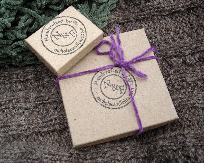 May include: Two brown cardboard boxes with a purple ribbon tied around the larger box. Both boxes have a round stamp with the text "Handcrafted by N&F nicholasandfelice.com".