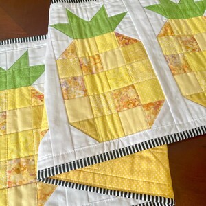 May include: Handmade quilted table runner featuring a repeating pineapple design. The pineapples are made of yellow and orange fabric squares with green tops. The runner has a white border with a black and white striped edge.