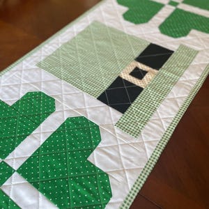 May include: A quilted table runner featuring a St. Patrick's Day theme. The design includes green shamrocks, a leprechaun hat, and a white background with a diamond pattern. The runner is trimmed with a green and white gingham border.