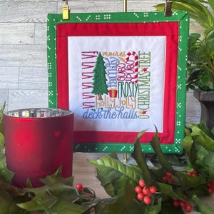 May include: A red and green Christmas-themed embroidered wall hanging with a white background. The embroidery features a Christmas tree, presents, candy canes, and the words "family", "movies", "cocoa", "frosty", "holly jolly", and "deck the halls".
