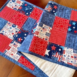 May include: Two patriotic table runners with a patchwork design. The runners feature red, white, and blue fabrics with star and stripe patterns, and text that reads "USA" and "Freedom". The edges are trimmed with a blue fabric.