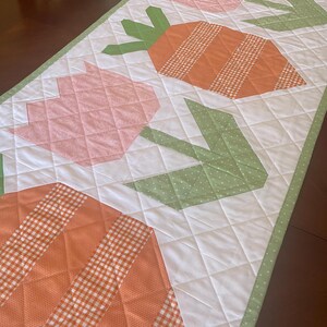 May include: A quilted table runner featuring a repeating pattern of orange carrots and pink tulips with green leaves. The carrots have a checkered pattern, and the leaves have a polka dot design. The runner is bordered with a green dotted fabric.