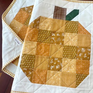 May include: Two quilted fabric mats featuring a patchwork pumpkin design in shades of mustard, gold, and cream. Each mat has a white background, a brown plaid stem, a green leaf, and a yellow and white gingham border with a small white label.