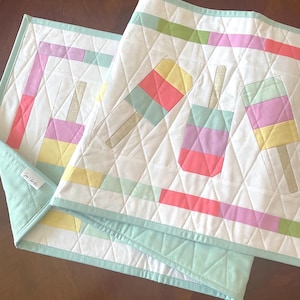 May include: A quilted table runner featuring a white background with a repeating pattern of colorful popsicle motifs in pastel yellow, pink, green, and blue. The runner has a light mint green border and diamond-shaped stitching throughout the fabric.