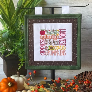 May include: A square fabric wall hanging featuring embroidered autumn-themed words including apples, foliage, autumn, spice, crisp air, corn, and pumpkins, with a red apple and sunflower icon, framed by a brown dotted border and a green outer border.