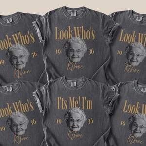 May include: A charcoal gray t-shirt featuring a vintage-style graphic of an elderly person's face. The text reads Look Who's 19 36 Khoe and It's Me! I'm 19 36 Khoe in gold lettering. The design includes a small inset photo box labeled Upload Photo.