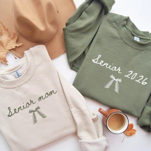 Puede incluir: Dos sudaderas, una crema y otra verde oliva, se muestran con un sombrero marrón y hojas de otoño. La sudadera crema dice "Senior mom", la verde dice "Senior 2026". Ambas tienen un gráfico de lazo. También se ve una taza de té.