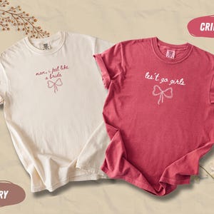 Op de afbeelding: Twee T-shirts, &eacute;&eacute;n ivoor en &eacute;&eacute;n karmozijnrood, worden getoond. Het ivoorkleurige shirt heeft de tekst "man, I feel like a bride" met een strik-afbeelding. Het karmozijnrode shirt zegt "les't go girls" met een strik-afbeelding. Beide zijn korte mouwen.