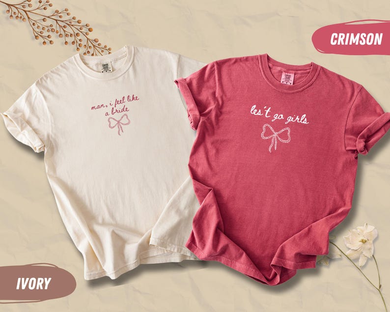 Op de afbeelding: Twee T-shirts, &eacute;&eacute;n ivoor en &eacute;&eacute;n karmozijnrood, worden getoond. Het ivoorkleurige shirt heeft de tekst "man, I feel like a bride" met een strik-afbeelding. Het karmozijnrode shirt zegt "les't go girls" met een strik-afbeelding. Beide zijn korte mouwen.