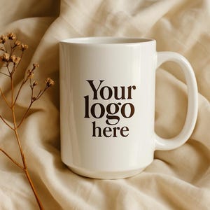 Puede incluir: Una taza de cerámica blanca con un asa grande, con el texto "Your logo here" en marrón. La taza está sobre un fondo de tela beige, con una ramita de flores secas a la izquierda.