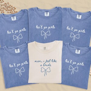 Op de afbeelding: Zes T-shirts, vijf in verbleekt blauw en &eacute;&eacute;n in cr&egrave;me. De blauwe shirts hebben de tekst "les't go girls" met een strik-afbeelding. Het cr&egrave;mekleurige shirt zegt "man, I feel like a bride" met een strik-afbeelding.