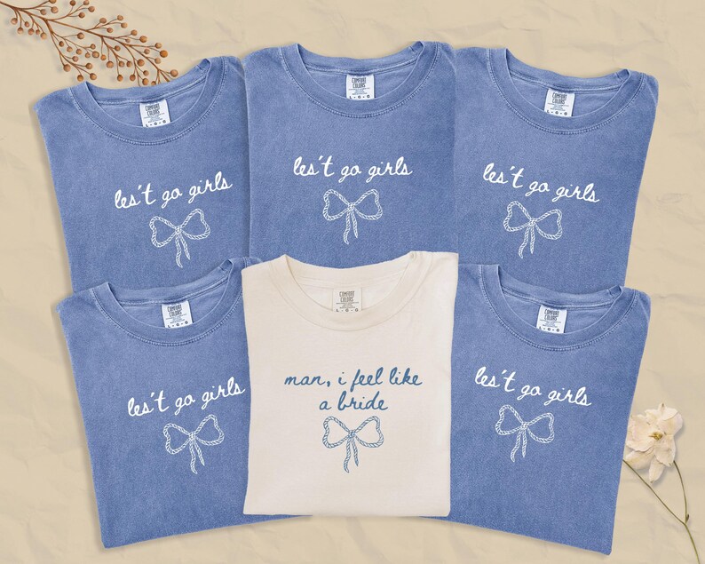 Op de afbeelding: Zes T-shirts, vijf in verbleekt blauw en &eacute;&eacute;n in cr&egrave;me. De blauwe shirts hebben de tekst "les't go girls" met een strik-afbeelding. Het cr&egrave;mekleurige shirt zegt "man, I feel like a bride" met een strik-afbeelding.