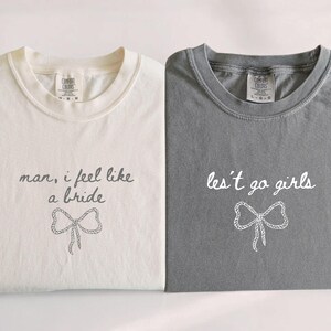 Op de afbeelding: Twee opgevouwen t-shirts, &eacute;&eacute;n cr&egrave;me en &eacute;&eacute;n grijs. Het cr&egrave;mekleurige shirt heeft de tekst "man, i feel like a bride" met een strik-afbeelding. Het grijze shirt zegt "les't go girls" met een strikafbeelding. Beide shirts hebben een ronde hals.