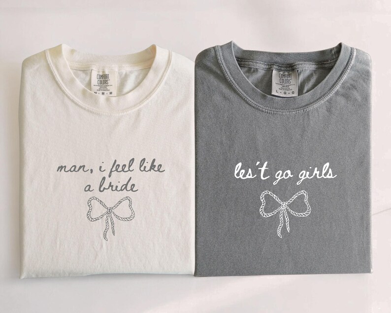 Op de afbeelding: Twee opgevouwen t-shirts, &eacute;&eacute;n cr&egrave;me en &eacute;&eacute;n grijs. Het cr&egrave;mekleurige shirt heeft de tekst "man, i feel like a bride" met een strik-afbeelding. Het grijze shirt zegt "les't go girls" met een strikafbeelding. Beide shirts hebben een ronde hals.
