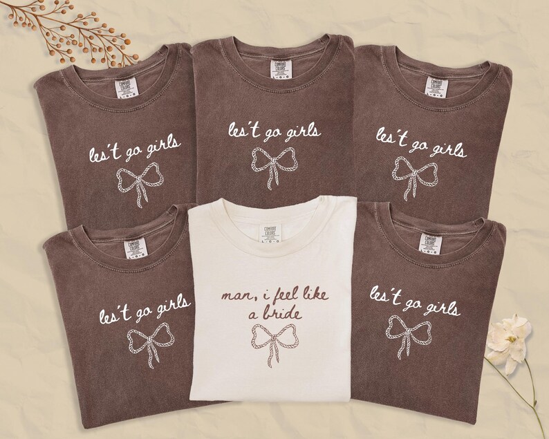 Op de afbeelding: Een verzameling bruine en witte t-shirts. De bruine shirts hebben de tekst "les't go girls" met een strik-afbeelding. Het witte shirt zegt "man, I feel like a bride" met een strik-afbeelding. Alle shirts hebben een ronde hals.