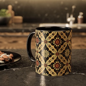 Puede incluir: Una taza de café negra con un diseño floral decorativo en dorado, negro y rojo. La taza tiene un asa y un interior negros, colocada sobre una encimera oscura. Ideal para café o té.