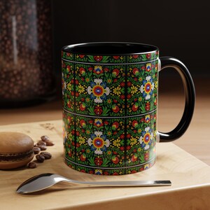 May include: A black coffee mug with a colorful floral pattern in red, blue, yellow, and white on a green background. The mug has a black interior and handle. A spoon and a macaron are in the foreground.