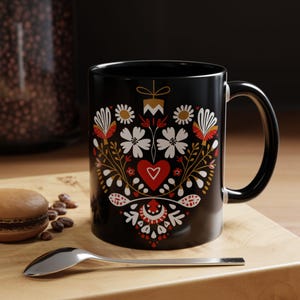 Può includere: Tazza in ceramica nera con un design floreale colorato. Il design include fiori bianchi, dettagli rossi e dorati. Un cucchiaio e un macaron sono su una superficie di legno.