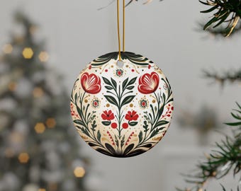 May include: Round Christmas ornament with a floral design. The ornament features red flowers, green leaves, and gold accents on a white background. It is hanging from a gold string, perfect for holiday decor.