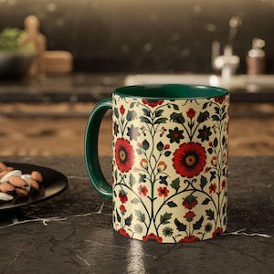 Op de afbeelding: Een keramische mok met een groene handgreep en binnenkant. De buitenkant heeft een bloemenpatroon met rode bloemen, zwarte accenten en groene bladeren op een crèmekleurige achtergrond. De mok is cilindrisch en staat op een donker oppervlak.