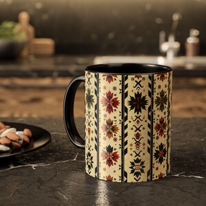 Puede incluir: Una taza de café con asa negra, fondo crema y un estampado floral repetido en rojo, verde y negro. El interior de la taza es negro.