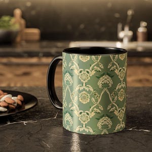 Puede incluir: Una taza de café de cerámica con asa e interior negros. El exterior presenta un estampado floral repetido en tonos verdes y crema sobre un fondo verde. La taza es cilíndrica y se apoya sobre una superficie oscura.
