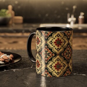 Puede incluir: Taza de café negra con un patrón floral decorativo en dorado, rojo y negro. La taza tiene un asa y un interior negros. El diseño recuerda a los azulejos tradicionales.