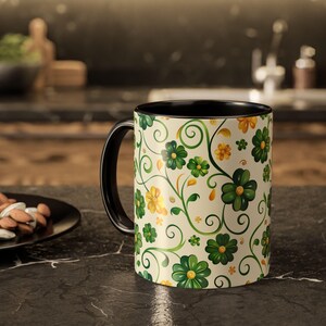 Puede incluir: Una taza de café de cerámica con asa e interior negros. El exterior presenta un estampado floral con flores verdes y amarillas, vides verdes y espirales sobre un fondo crema. La taza está sobre una encimera oscura.