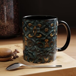 Puede incluir: Una taza de cerámica con interior y asa negros. El exterior presenta un diseño en relieve verde azulado y dorado con motivos florales y de cinta. La taza está sobre una superficie de madera con una cuchara, granos de café y un macaron.