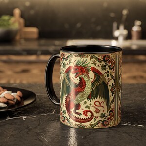 Puede incluir: Una taza de cerámica con asa e interior negros. La taza presenta una ilustración detallada de un dragón rojo y verde con las alas extendidas, sobre un fondo crema con detalles florales. El diseño del dragón está enmarcado por un borde decorativo.