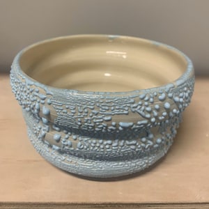 May include: A light blue ceramic bowl with a textured, raised design. The bowl has a white interior and a light blue exterior.
