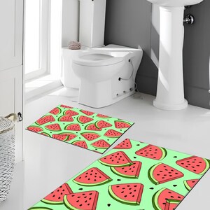 Op de afbeelding: Twee rechthoekige badmatten met een patroon van watermeloen schijfjes. De schijfjes zijn rood met zwarte zaden en groene contouren, op een lichtgroene achtergrond. In een badkamer met een witte toilet en wastafel.