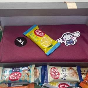 Könnte beinhalten: Eine graue Box mit rotem und weißem gemustertem Seidenpapier im Inneren. Die Box ist gefüllt mit verschiedenen Geschmacksrichtungen von Kit Kat Schokoladenriegeln, darunter Milchschokolade, Tee, Eiscreme und Salz-Zitrone. Die Box enthält auch einen kleinen Aufkleber mit einem Comic-Charakter mit Hasenohren.
