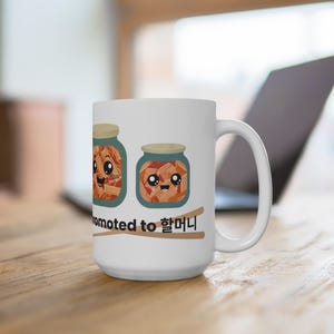 May include: White ceramic mug featuring two cartoon jars of kimchi with smiling faces. The text "Promoted to 할머니" is printed below the jars. The mug has a large handle and is set on a wooden surface.
