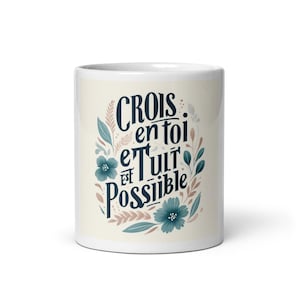Peut inclure: Un mug en céramique blanc avec l'inscription française "Crois en toi et tout est possible" en écriture bleu marine. Le design comprend des fleurs et des feuilles bleu sarcelle, et du feuillage beige et rose.