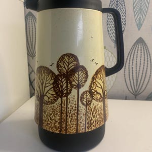 Op de afbeelding: Een vintage geïsoleerde thermoskan met een zwarte deksel, handvat en basis. De crèmekleurige behuizing heeft een bruin boom- en vogelontwerp. De thermoskan is ontworpen om dranken warm of koud te houden.