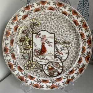 Könnte beinhalten: Ein Satz dekorativer Teller mit Blumen- und Vogelmuster. Die Teller haben einen weißen Hintergrund mit einem braunen und orangefarbenen Blumenrand. Die Mitte der Teller zeigt einen Hahn, Vögel und Blumenillustrationen.